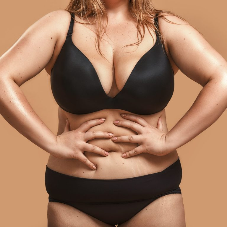 I love my body. Plump happy plus size woman in sexy black lingerie touching her belly while standing against brown background. Studio portrait. Overweight female